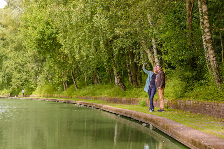 Un homme et une femme se baladant sur un chemin entre forêt et cours d'eau et se prenant en photo avec un smartphone © WBT - J.P. Remy